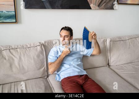 Guy coping with summer heat lying on sofa using paper fan suffocating of stuffiness, sweltering heat Stock Photo