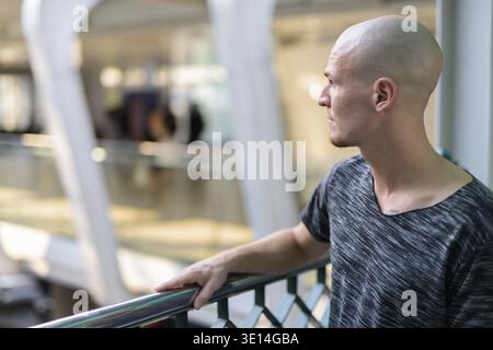 Profile view of young handsome bald man thinking on the foot bridge against view of the street and exterior of mall in the city Stock Photo