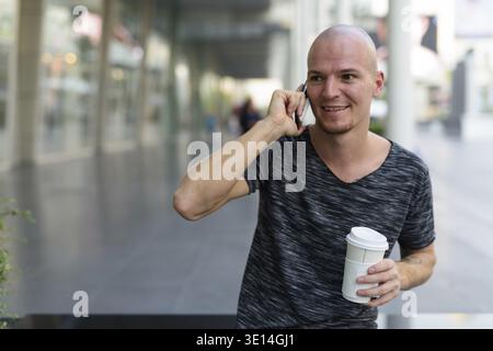 Thoughtful young happy bald man smiling and holding paper coffee cup while talking on mobile phone outside of the mall in Bangkok Thailand Stock Photo