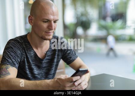 Young handsome bald man using mobile phone inside restaurant with glass windows Stock Photo