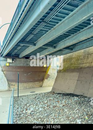 People walk along a path under a large metal bridge. Concrete walls and rocks are visible on either side. Sunlight comes through from above while shad Stock Photo