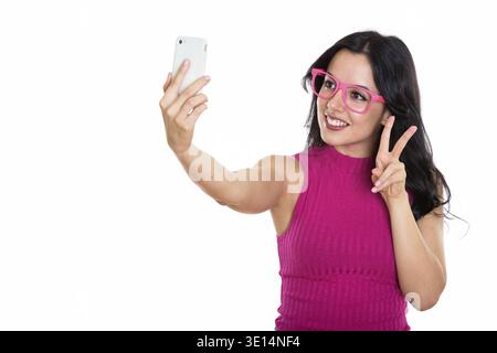 Studio shot of young happy Spanish woman smiling while taking selfie picture with mobile phone and giving peace sign Stock Photo