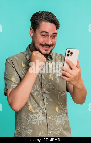 Middle-aged man looks at smartphone triumphantly clenches fist in joyful reaction, celebrating success or great news. Young adult guy isolated on blue background shows happiness and energetic emotion Stock Photo