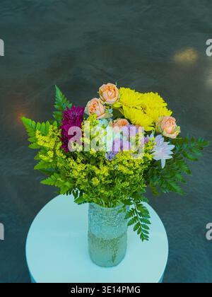 A mixture of colorful flowers is arranged in a transparent vase placed on a round table. The setting is bright and features a smooth surface reflectin Stock Photo