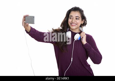 Young happy Indian woman smiling while wearing headphones around neck and taking selfie picture with mobile phone Stock Photo