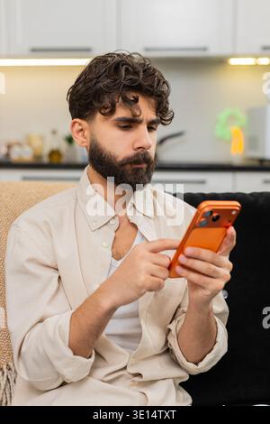 Indian man at home works on smartphone typing messages for freelance clients in chat app busy at work. Arabian guy checks orange mobile phone writes reply to customer message feels busy concentrated. Stock Photo