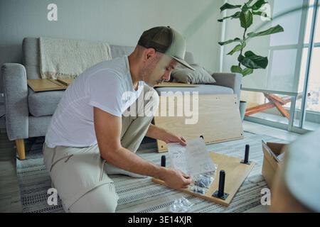 Man kneeling on floor reading an instruction manual while assembling furniture Stock Photo