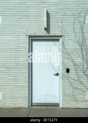 A white door stands on a light-colored brick wall. The shadow of a tree is visible on the wall, and the sidewalk is clear. The scene is empty and quie Stock Photo