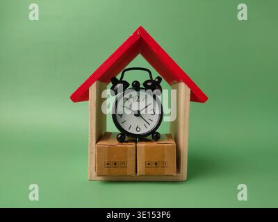 Small black alarm clock resting on two miniature cardboard shipping boxes in wooden house frame. Set against a solid green background. Stock Photo