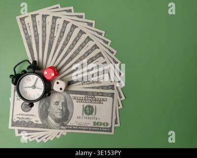 Small black alarm clock and two stacked dice resting on a fan of US dollar bills on a green background. Stock Photo