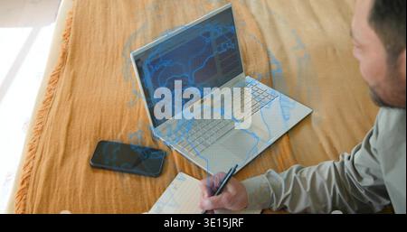 Writing mature man wearing gray shirt, leaning on bed, using silver laptop and notebook, copy space Stock Photo