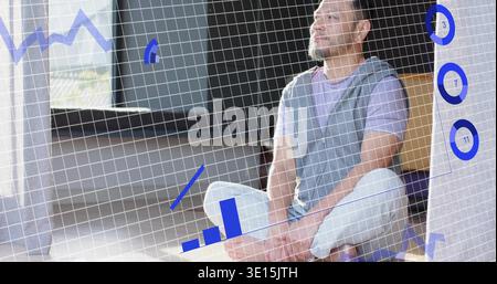 Sitting mature Asian man wearing tee and vest gazing upward on cushion showing blue data overlays Stock Photo