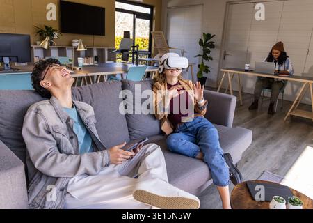 Diverse coworkers sitting on sofa using white VR headset while man holding phone in shared office Stock Photo