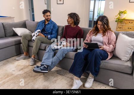 Diverse friends sitting on gray sectional sofa at home holding VR controllers, headset and tablet Stock Photo