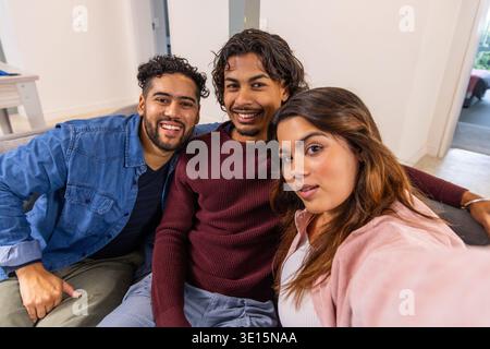 Diverse friends leaning in, posing for selfie on living room couch with smartphone and cushions. Trio, group, candid, casual, home, interior, daylight Stock Photo