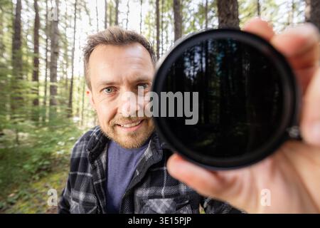 Man smiles through a neutral density filter for the camera . Green trees blur behind his face. Natural light filters through the canopy. He holds the Stock Photo