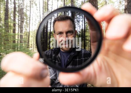 Man smiles through a neutral density filter for the camera . Green trees blur behind his face. Natural light filters through the canopy. He holds the Stock Photo
