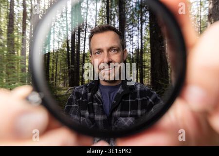 Man smiles through a neutral density filter for the camera . Green trees blur behind his face. Natural light filters through the canopy. He holds the Stock Photo
