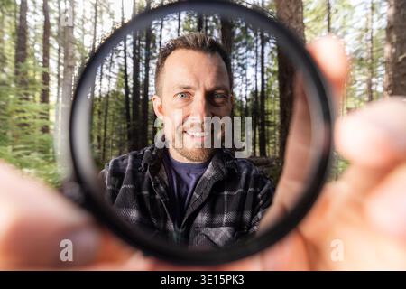 Man smiles through a neutral density filter for the camera . Green trees blur behind his face. Natural light filters through the canopy. He holds the Stock Photo