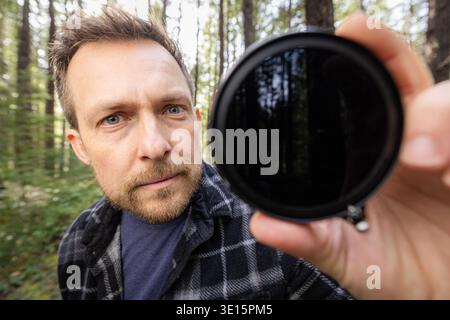 Man smiles through a neutral density filter for the camera . Green trees blur behind his face. Natural light filters through the canopy. He holds the Stock Photo