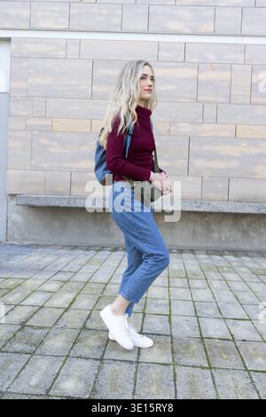Female pausing beside beige tiled wall on paved plaza, carrying blue backpack and green bag Stock Photo