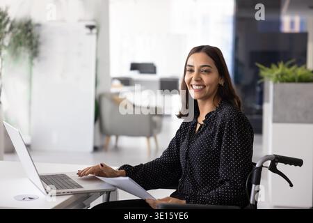 Positive Middle Eastern female worker in wheelchair using laptop computer Stock Photo