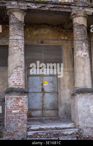 weathered neoclassical facade featuring crumbling brick pillars and a rusted industrial metal door in an abandoned building with a gritty atmospheric Stock Photo