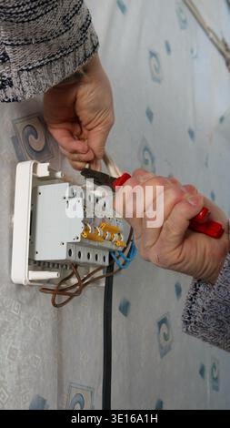 A man's hands hold pliers and secure the wires going to a wall-mounted home electrical distribution box with circuit breakers. Stock Photo