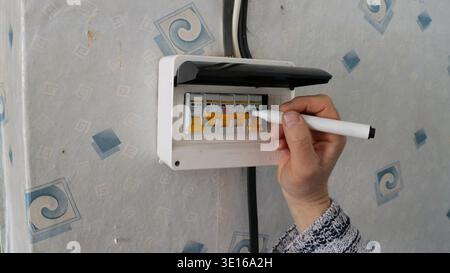 A hand holding a black marker applies labels to the circuit breakers in a residential distribution board, and also marks the meter mounted on the wall Stock Photo