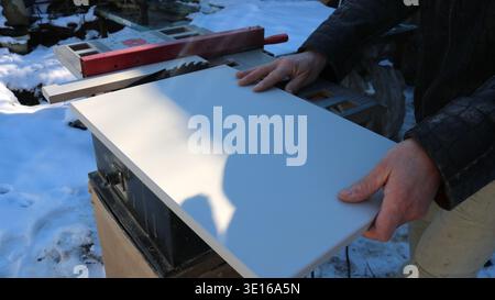 In an open woodworking shop, a carpenter presses a compressed board against the work surface of a circular saw to cut the workpiece, using an end saw Stock Photo