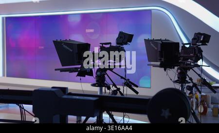 Professional broadcasting cameras on tripods and rails capturing content in a modern television studio with a large screen displaying abstract visuals Stock Photo