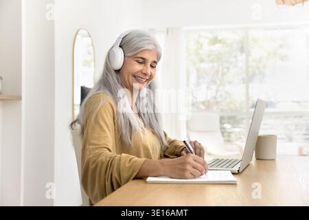 Older woman wearing headphones writing in notebook while using laptop Stock Photo