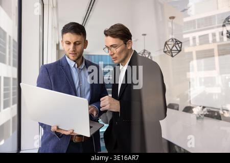 Two businessmen stand in boardroom, deeply focused on laptop screen Stock Photo