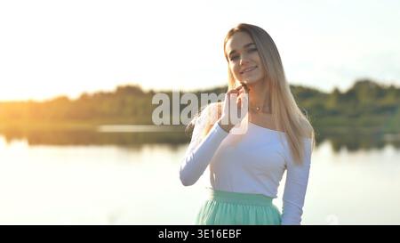 Young woman smiling, enjoying golden sunset hour by peaceful lake Stock Photo