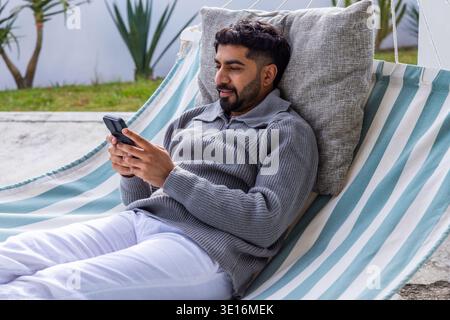 Man in mid-30s reclining on striped hammock on patio, holding smartphone and wearing gray pullover Stock Photo