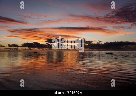 Colorful sunset sky reflecting on calm ocean water with dark clouds and small boats on horizon. Stock Photo
