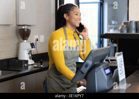 African American woman barista wearing gray apron speaking on phone behind counter by POS terminal Stock Photo