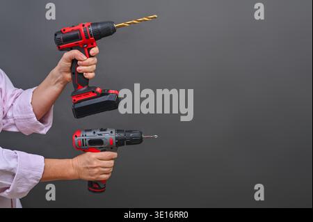 Two cordless drills raised in hands, red and black power tools with extended bits, isolated on neutral background, Hands holding two drills, modern co Stock Photo
