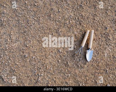 Trowel and hand fork placed on dry soil in a garden area with gravel and dirt visible Stock Photo