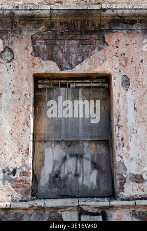 Old abandoned building with boarded-up window. Cracked wall with peeling plaster and exposed red bricks.  Boarded window on an old weathered wall. Con Stock Photo