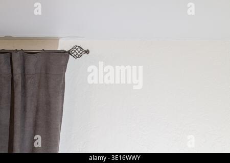 A detailed view of a dark gray fabric curtain partially drawn on a decorative metal curtain rod, set against a white textured wall Stock Photo