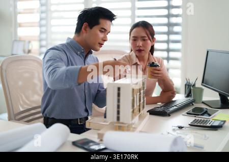 Two Asian adult architects intently collaborate on a detailed building model in their bright contemporary office space developing new construction pla Stock Photo