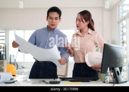 Professional Asian engineers collaborate reviewing architectural plans in a contemporary office setting discussing building project development Stock Photo