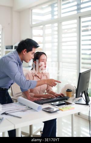 Asian business professionals analyze architectural blueprints on a desktop computer. Focused discussion and collaborative problem solving in a modern Stock Photo