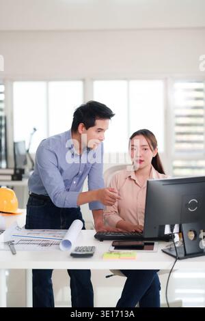 Asian adult professionals collaborate at office desk. Man points to computer screen. Woman types. Teamwork on architectural blueprints and design. Pro Stock Photo