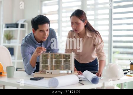 Two professional Asian architects intently discuss skyscraper design plans. They collaboratively examine an architectural model within a modern bright Stock Photo