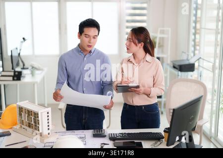 Professional Asian design team reviewing building blueprints in modern office space for new construction project planning successful urban development Stock Photo