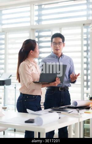 Adult Asian coworkers reviewing architectural blueprints on laptop discussing innovative design ideas in bright contemporary office Stock Photo