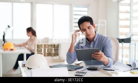 Adult asian male architect thoughtfully examines architectural plans on a tablet computer in a bright contemporary workspace Focus vision development Stock Photo