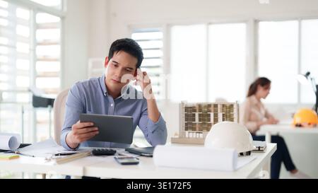 Adult Asian architect concentrating on project plans using tablet. Modern office. Blurred colleague works in background. Professional design focus. Stock Photo